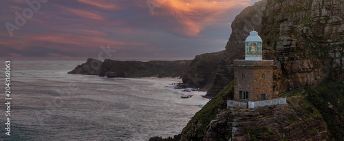 Fotografia A compact stone lighthouse with glass lantern stands on a rugged cliff as surf churns at dusk