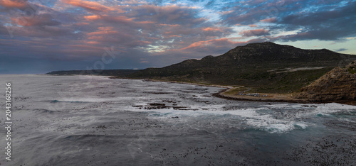 Rugged Cape of Good Hope curves into the Atlantic near Cape Town. Low mountains and fynbos meet rocky shore, foamy surf, and pastel sunset light on sea and clouds.