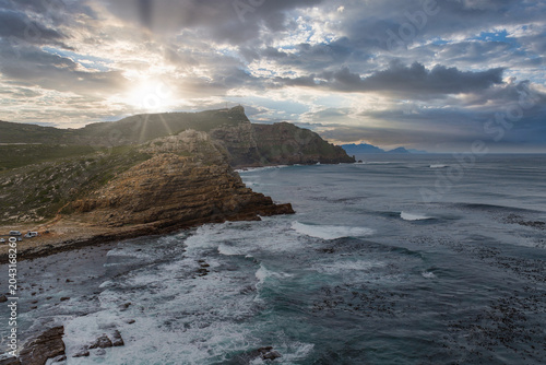 Rugged Cape of Good Hope near Cape Town, South Africa, shows layered cliffs, a long shoreline, white surf, and kelp beds as sunbeams filter through heavy clouds.
