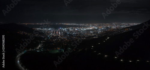 A wide city basin view shows Cape Town at night from mountain slopes, with a curving road of headlights, tall lit buildings, and suburbs extending under an overcast sky.