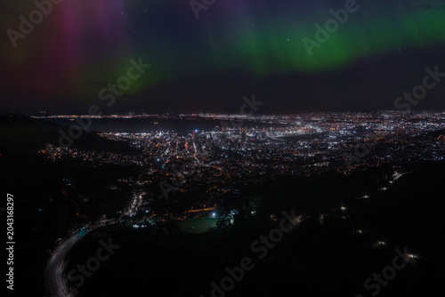 Tableau sur toile Aerial night scene over Cape Town shows city lights, Table Bay, and a winding mountain road, as green and magenta auroral ribbons sweep the sky in long exposure