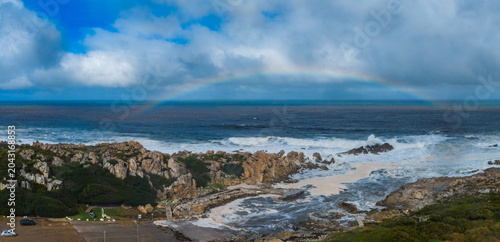 Tablou pe pânză Rugged Cape of Good Hope near Cape Town, South Africa, shows jagged granite, fynbos slopes, and foaming surf as a faint rainbow arcs under thick storm clouds
