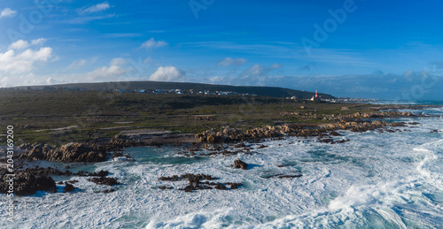 Aerial view shows Cape Agulhas Lighthouse on a low peninsula in South Africa as surf breaks over rocky outcrops, with a sparse settlement under a clear daylight sky.