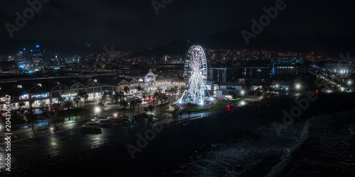 Night view of Cape Town V and A Waterfront with the Cape Wheel lit, wet streets reflecting lights, harborfront and promenade glow, skyline and Table Mountain recede.