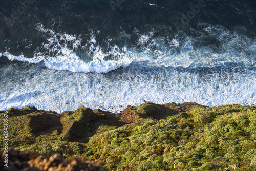 Cuadro en lienzo High angle shot looking down at the blue ocean waves breaking into white foam against a rocky cliff covered in green plants and shrubs during daylight