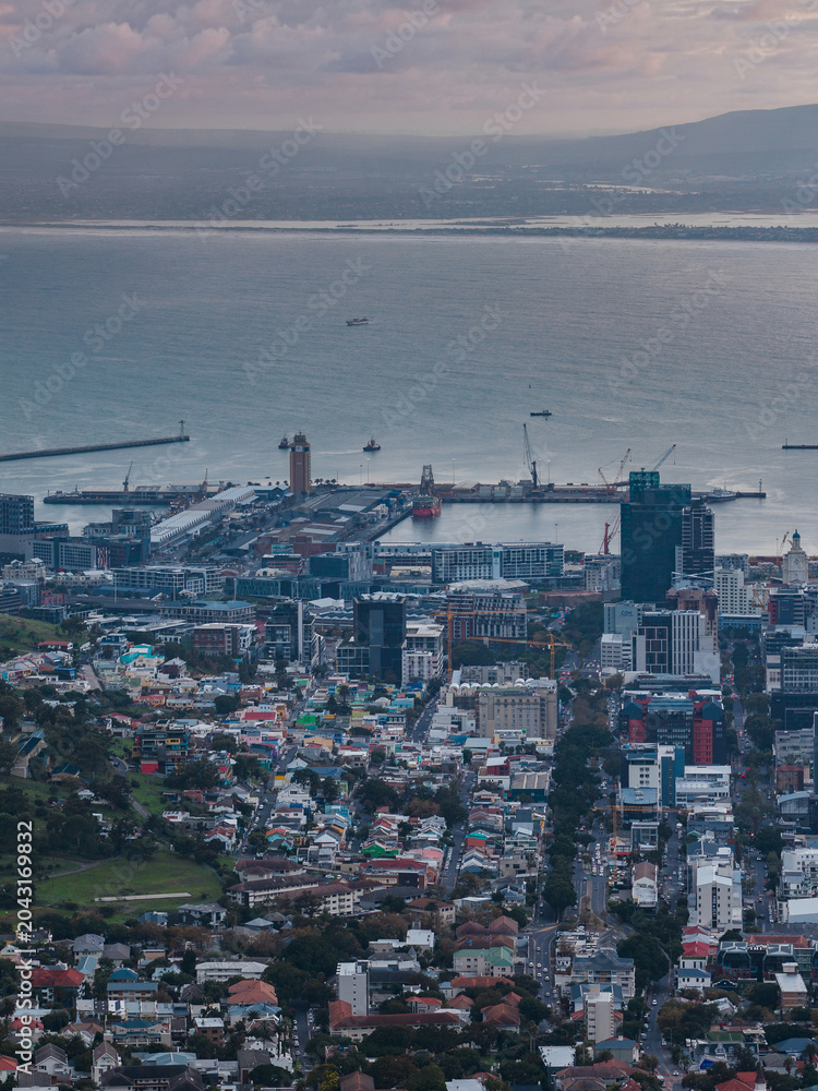 Obraz premium Aerial view of Cape Town city center and Victoria and Alfred Waterfront shows piers, cranes, vessels, mid and high rises, homes, parkland, dusk light, mountains, clouds.