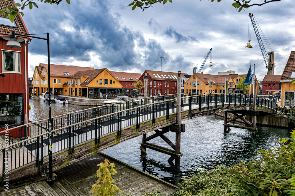 Fototapeta premium Scenic waterfront canal with colorful historic buildings and moored boats under dramatic sky, Kristiansand, Norway