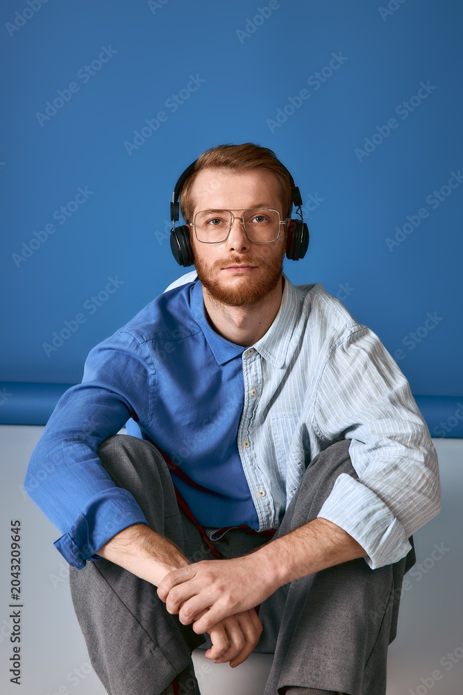 Fototapeta premium Handsome red haired man in stylish attire enjoys music while sitting on a blue backdrop.
