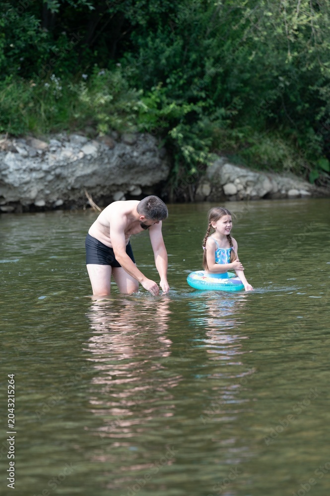 Fototapeta premium A father and daughter enjoy a summer day playing in a river. The girl is using a small inflatable ring.