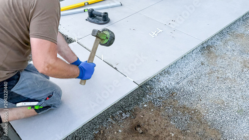Male worker laying large concrete tiles on gravel surface using a rubber mallet, tools and measuring equipment visible in the construction area