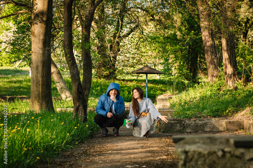 Fototapeta premium Happy couple crouching and posing on a path in a sunny spring park. Man and a woman enjoying nature together. Mom and dad are sitting and watching their children play off-screen.