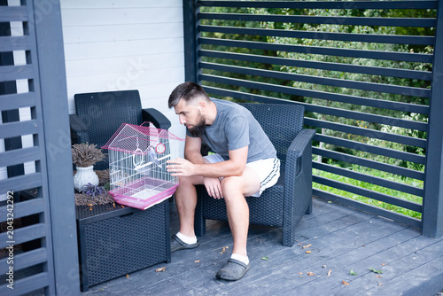 Man with beard sits outside, looking at a bird in a cage.