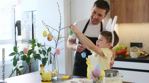 Father and young son hang colorful eggs on a decorative tree in a contemporary kitchen, surrounded by Easter-themed decorations and spring flowers