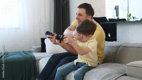 Father and young boy engaged in video gaming on a couch, with a contemporary kitchen area and dining chairs in the background