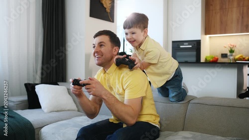 Father and young boy engaged in video gaming on a couch, with a contemporary kitchen area and dining chairs in the background
