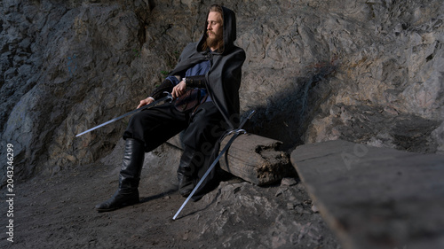 Foto A hooded warrior with swords rests on a rocky outcrop, exuding a mysterious and powerful aura