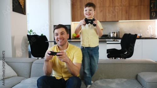 Father and son playing video games together on a couch in a modern living room with a kitchen visible in the background
