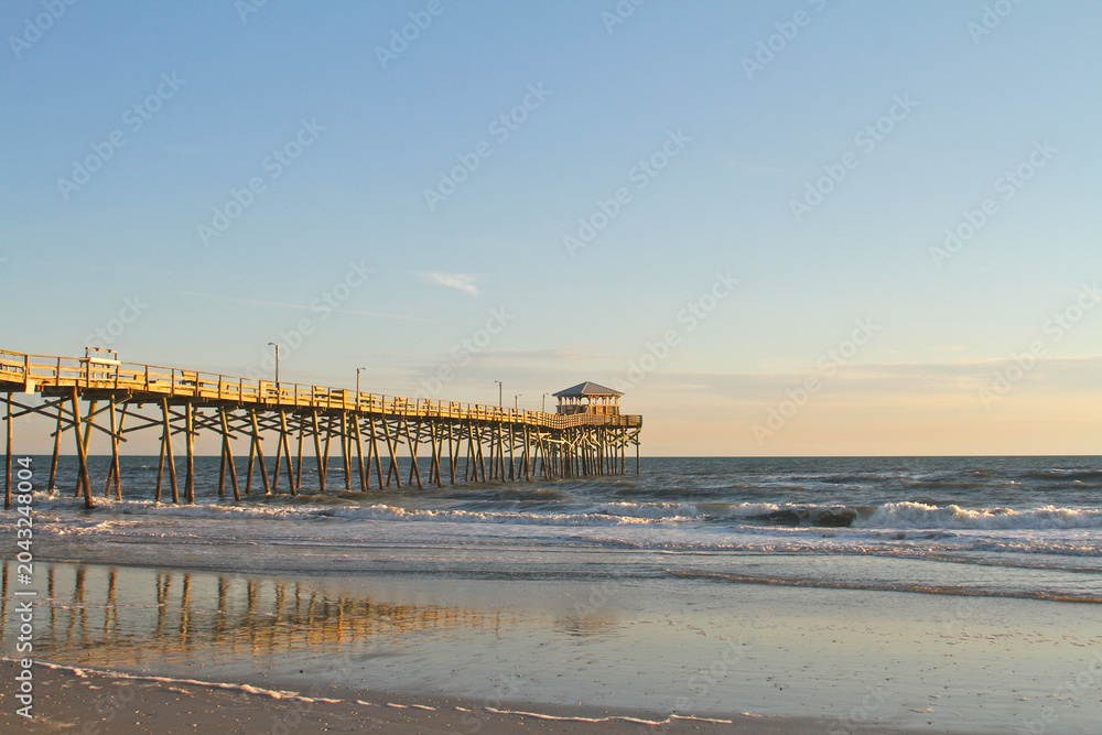Fototapeta premium The Atlantic Beach Fishing Pier extends into the Atlantic Ocean as evening light reflects across wet sand and rolling waves along the North Carolina coast.