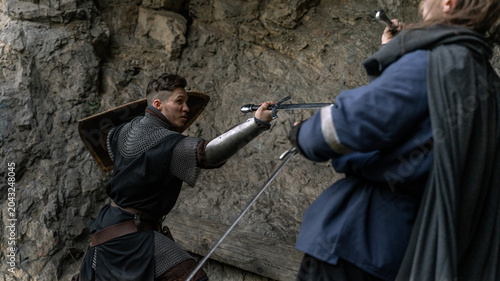 Photos A determined warrior, clad in armor and wielding a sword, confronts an opponent amidst a rugged stone landscape