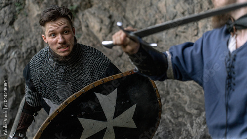 Tableau sur toile A medieval knight with a shield and chainmail faces off against an opponent wielding a sword during a dramatic battle scene