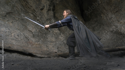 Bilde på lerret A rugged warrior stands poised with a gleaming sword amidst a rocky cavern, dust swirling around his feet