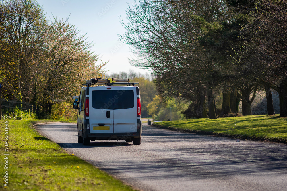 Fototapeta premium Van Driving on Curving Suburban Road Through Green Landscape on Sunny Afternoon in England UK