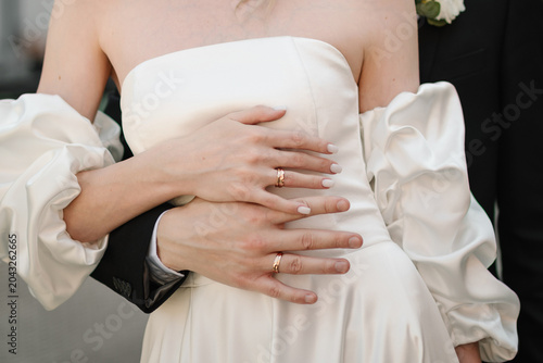 Romantic wedding detail. Closeup of a newlywed couple during an intimate embrace. The groom stands behind the bride and gently holds her waist with hands displaying their gold wedding rings.