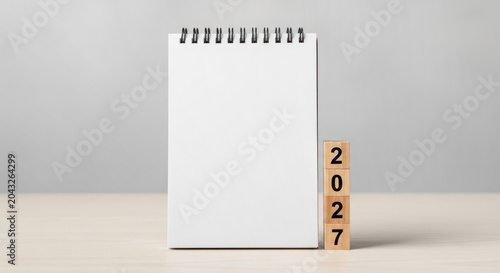 Blank spiral notepad with wooden blocks displaying the year 2027 on a light wooden table against a soft gray background