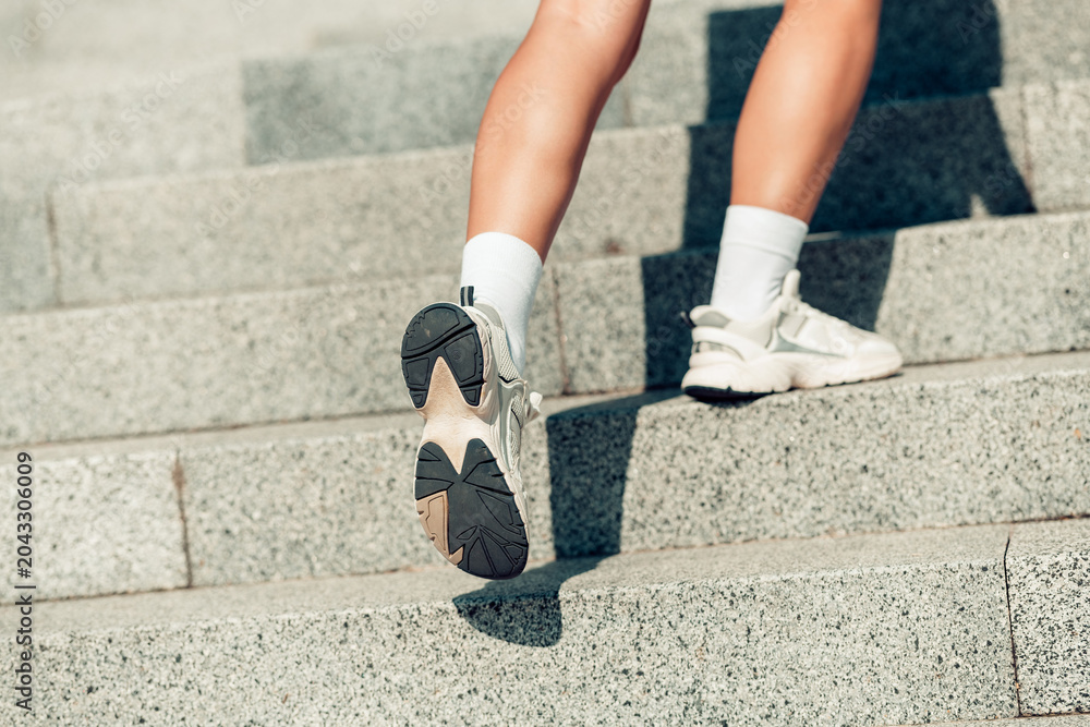 Naklejka premium Woman walking up stone stairs in sneakers for fitness and urban workout, close-up of legs and shoe sole movement on outdoor steps