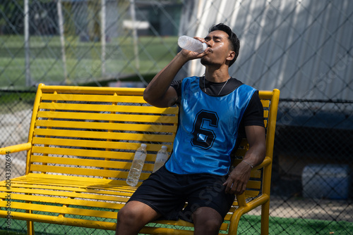 A Thai Asian soccer player drinks from a water bottle while resting on a yellow bench beside an outdoor field during a break from play