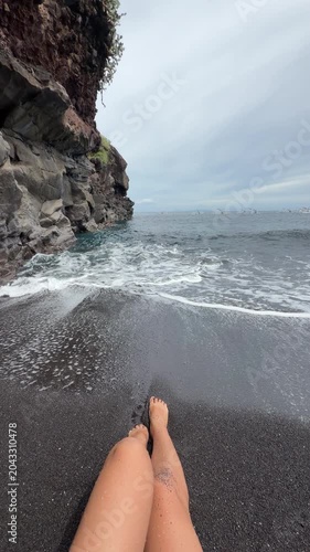Vertical POV of female legs in ocean waves on black sand beach 