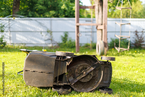 Fotografie An electric lawn mower lying upside down on green grass exposing the sharp cutti