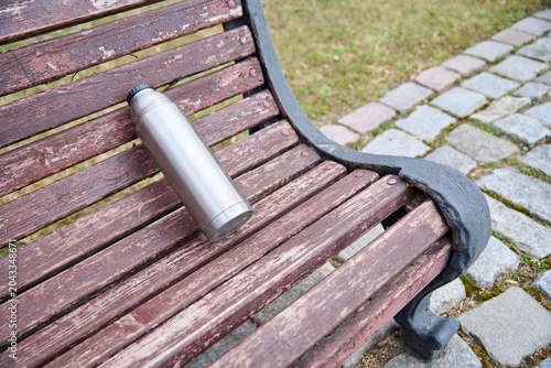 Картината върху платно Silver thermos bottle resting on weathered wooden park bench with stone path and