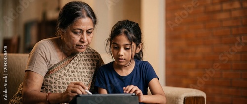 Indian grandmother granddaughter tablet learning together in living room, elderly woman teaching young South Asian girl using digital stylus device on sofa, family bonding and digital education concep