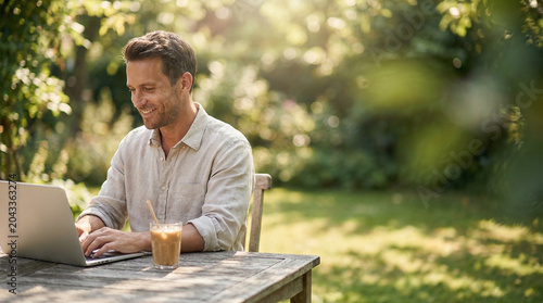 A smiling man in a linen shirt sitting at a wooden table in a green, sun-drenched garden, working on a laptop with an iced coffee. Illustrates the freedom of remote work.