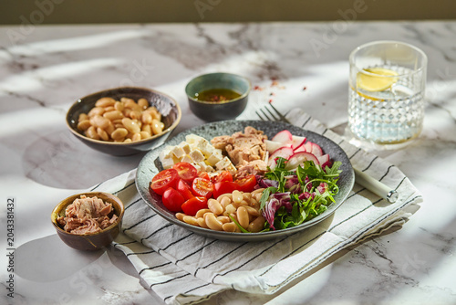 Tuna salad bowl with cannellini beans, feta cheese, cherry tomatoes, radish, and greens served on a marble table with olive oil dressing.