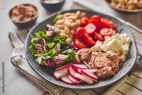 Top view of healthy tuna salad bowl with white beans, feta cheese, tomatoes, radish, arugula, and olive oil dressing on a marble surface.