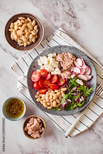 Top view of healthy tuna salad bowl with white beans, feta cheese, tomatoes, radish, arugula, and olive oil dressing on a marble surface. Top view