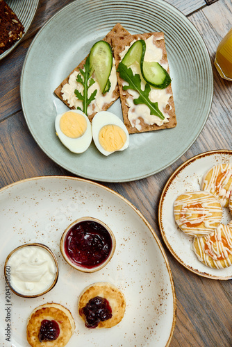 Delicious breakfast spread with cheese pancakes (syrniki), fried eggs, fresh salad, toasts, coffee and orange juice on wooden table. Healthy morning meal concept, variety of dishes.