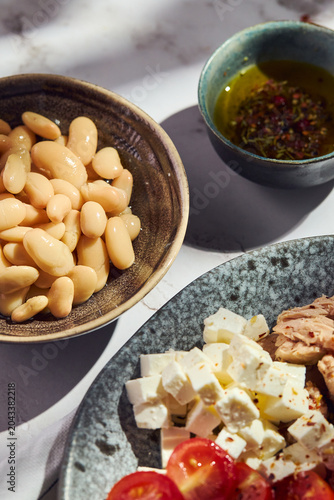 Top view of fresh ingredients for a healthy tuna and bean salad. Includes white beans, cubed feta cheese, cherry tomatoes, canned tuna, and spiced olive oil on a marble table.