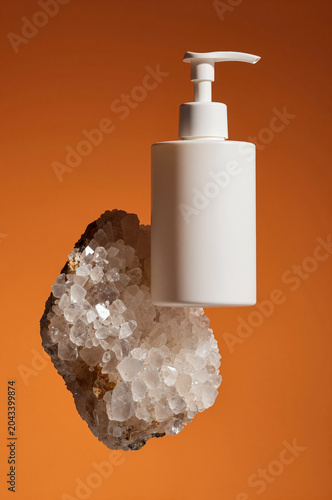 Cream dispenser floats above a jagged crystal cluster against an ochre backdrop. Soft side lighting highlights the matte plastic texture and translucent mineral facets.