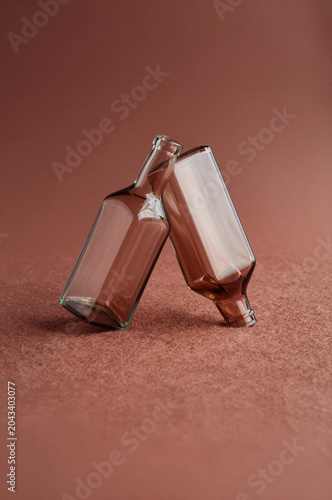 Glass bottles lean against one another on a textured surface under warm directional light. Two transparent containers rest at an angle amidst soft amber tones and subtle reflections.