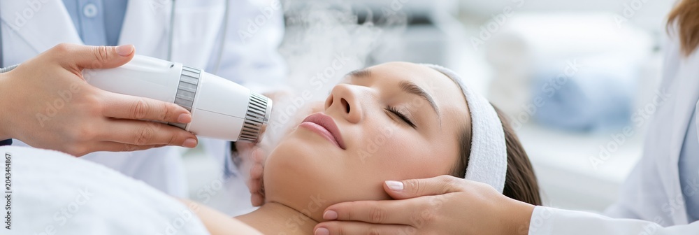 Fototapeta premium Crisp daylight highlights a facial technician using a vapor device in a bright clinical spa lab focusing on the hands and face while medical charts and towels remain unfocused