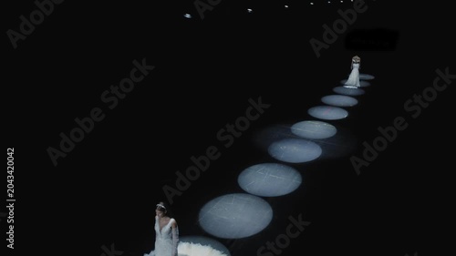 Cinematic wide shot of two elegant models in white wedding gowns standing on dark runway illuminated by circular LED ring lights at bridal fashion show, shot from elevated angle. Perfect for wedding a