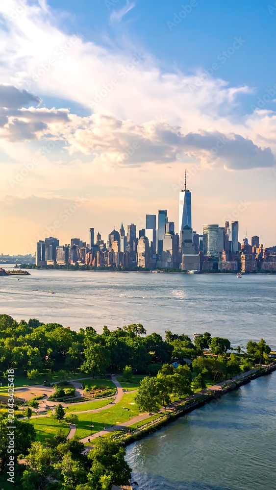 Fototapeta premium Sunlit Manhattan Skyline Rising Beyond River and Green Waterfront Park