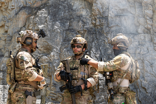 Group of three soldiers in camouflage uniform talking during military tactical briefing outdoor. Special forces operators preparing weapons and gear near rock cliff