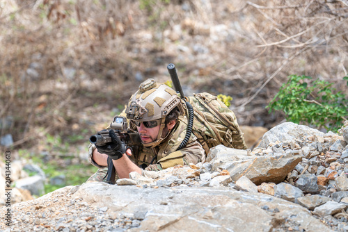 Billede på lærred Soldier in camouflage uniform lying in prone position aiming rifle behind rocks