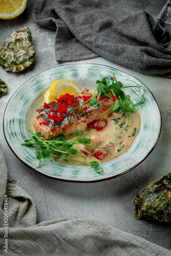 Vertical restaurant dish with grilled salmon fillet, creamy sauce, lemon, red caviar, tomatoes and fresh microgreens on ceramic plate