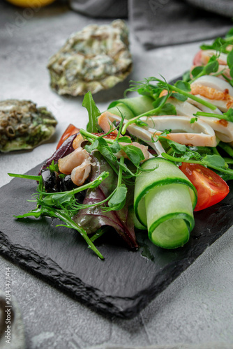 Vertical close-up of seafood salad with squid, cucumber ribbons, olives, tomatoes and fresh greens served on a black slate plate