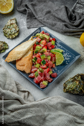 Vertical composition of tuna tartare with avocado, cucumber, tomatoes, lime, capers and toasted bread on a gray restaurant plate